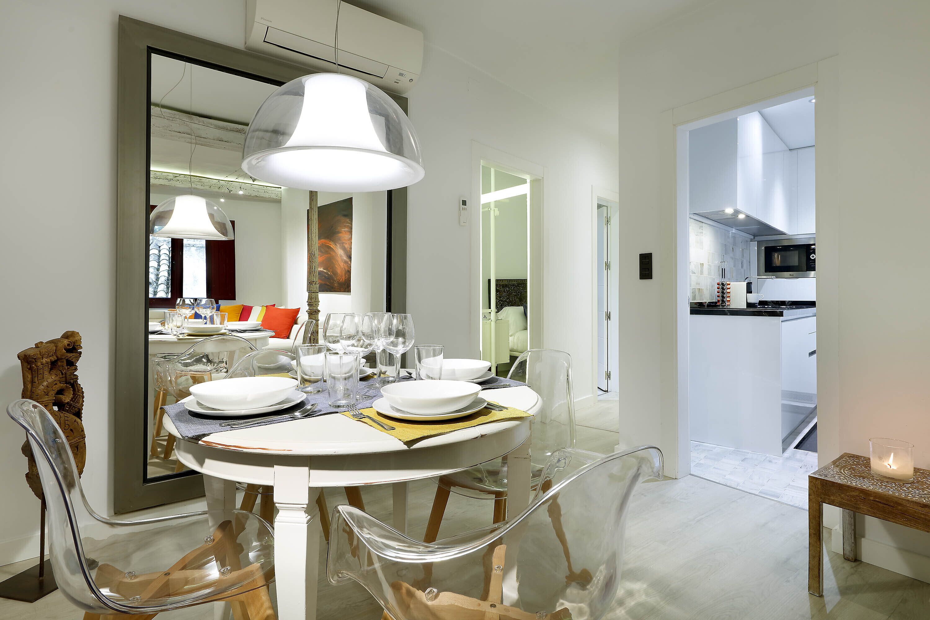 Een eetruimte met een ronde tafel voor vier personen, transparante stoelen en een hanglamp. Een keuken is zichtbaar door een open doorgang.