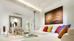 Een moderne woonkamer met een witte bank, kleurrijke kussens, een glazen salontafel en een groot abstract schilderij aan de muur.