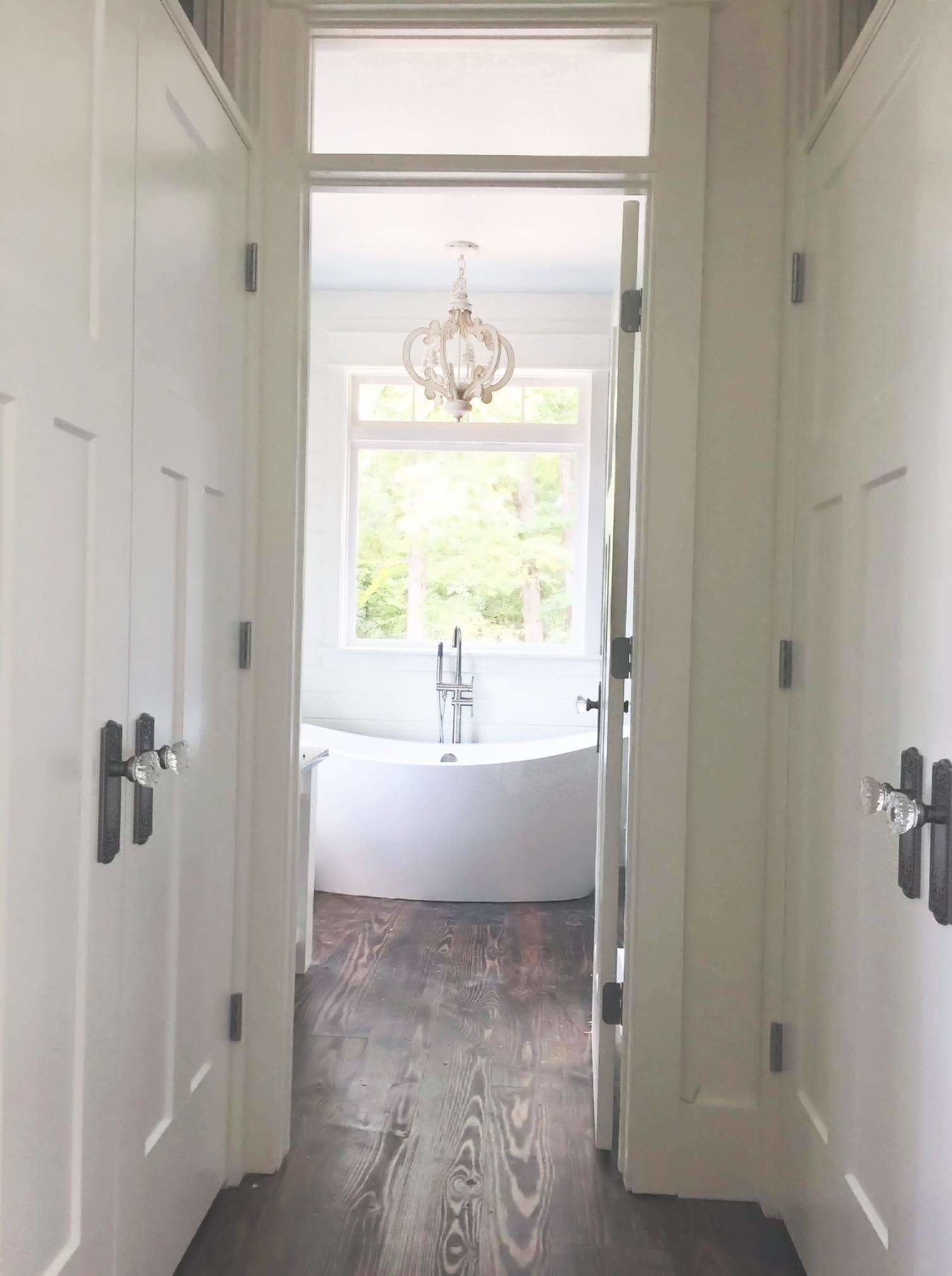 A hallway leading to a bathroom with a bathtub, a window, and a chandelier.