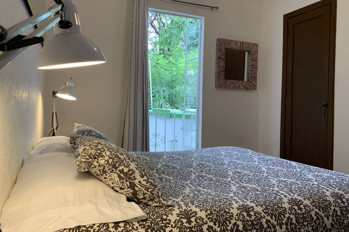 A bedroom with a bed, a bedside lamp, a window with curtains, a door, and a mirror on the wall.