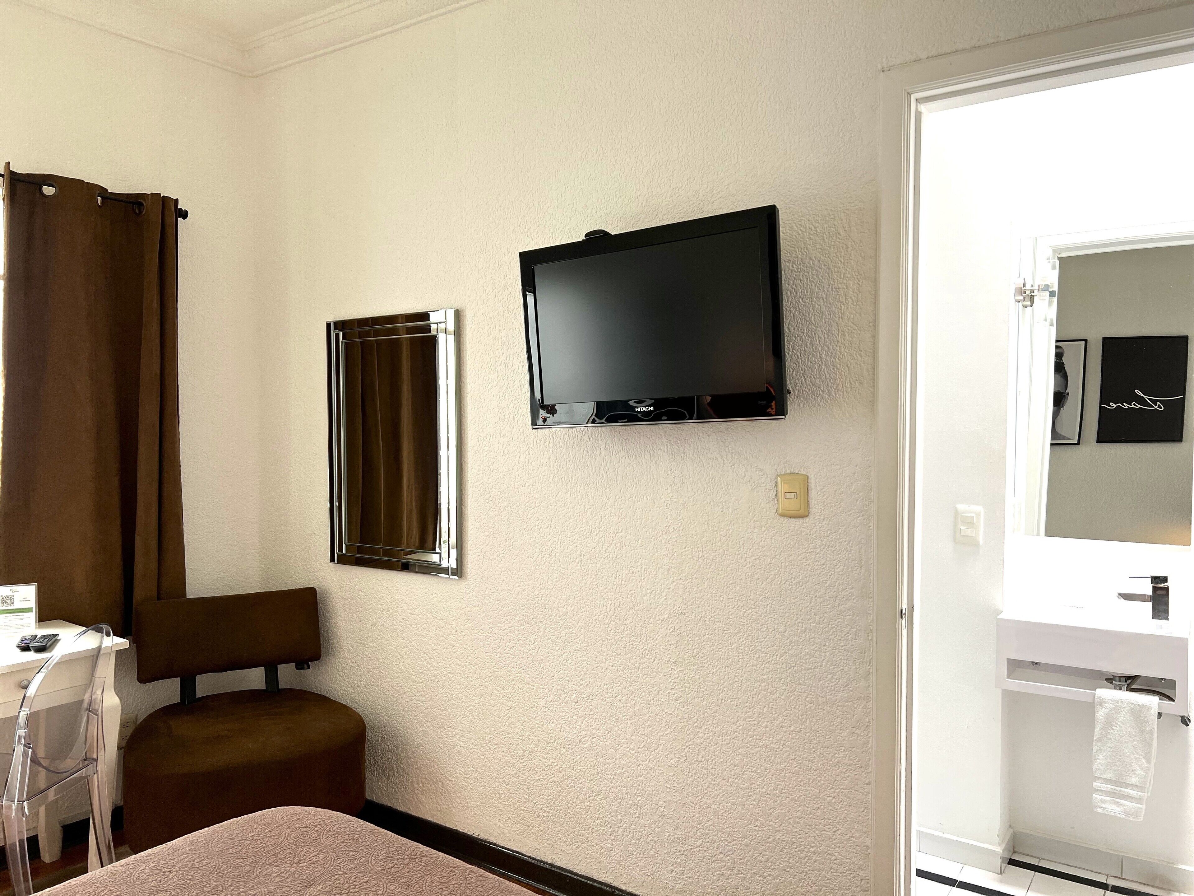 Habitación de hotel con cama, escritorio con silla, televisión montada en la pared, baño con espejo y perchero, y cortina en la ventana.
