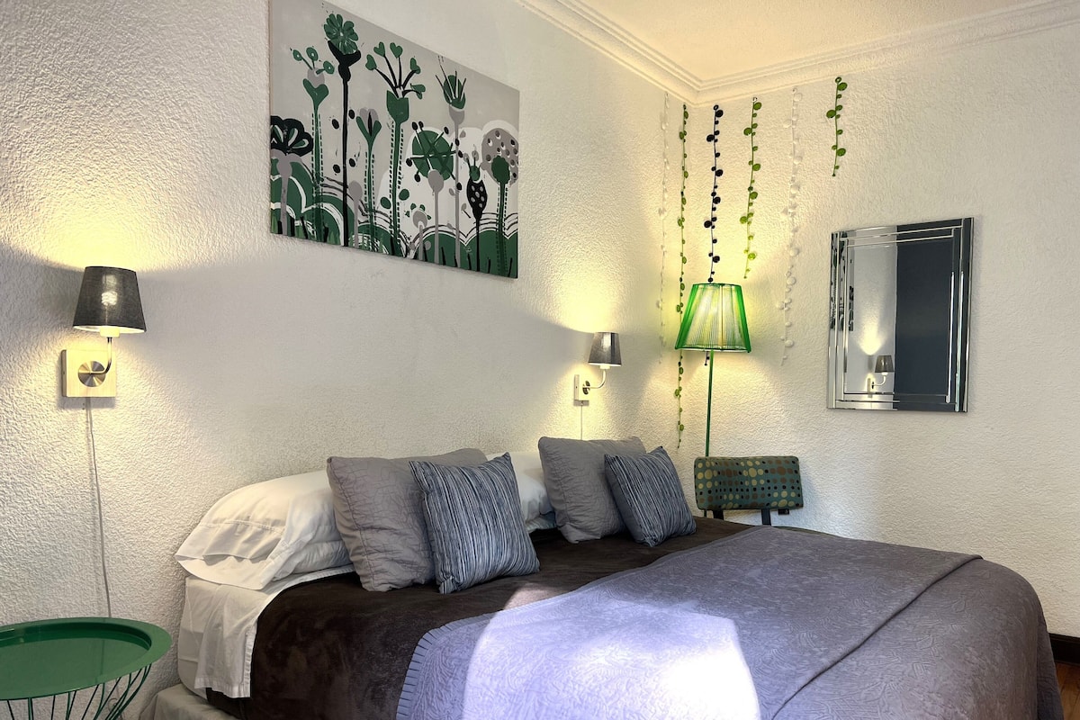 A bedroom with a large bed, multiple pillows, two wall-mounted lamps, a green chair, and a wall art piece.