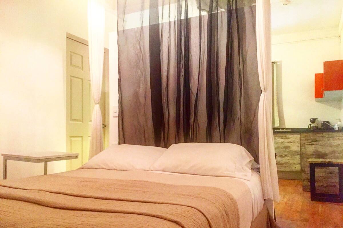 A bedroom with a bed, a bedside table, a curtain, and a view of a kitchen area.
