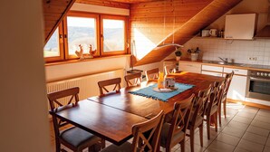 Een eetgedeelte met een houten tafel en stoelen, een keuken met witte tegels, en uitzicht op de bergen door grote ramen.