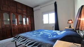 Een slaapkamer met een bed, een houten kledingkast, een raam met gordijnen, een tafel en een lamp.