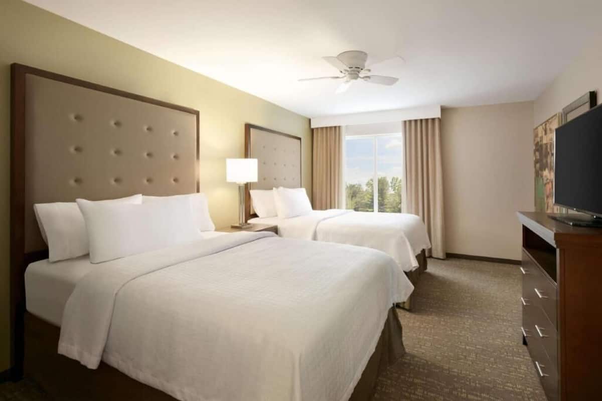 A hotel room with two beds, a ceiling fan, a TV, and a window with curtains.