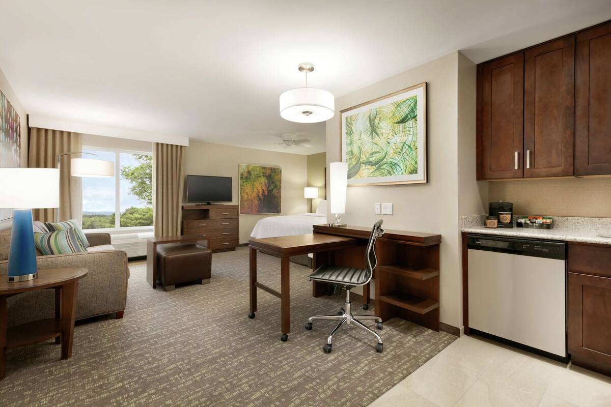 Een hotelkamer met een bank, een bureau, een stoel, een televisie, een schilderij en een keukenhoek met kastjes en een vaatwasser.