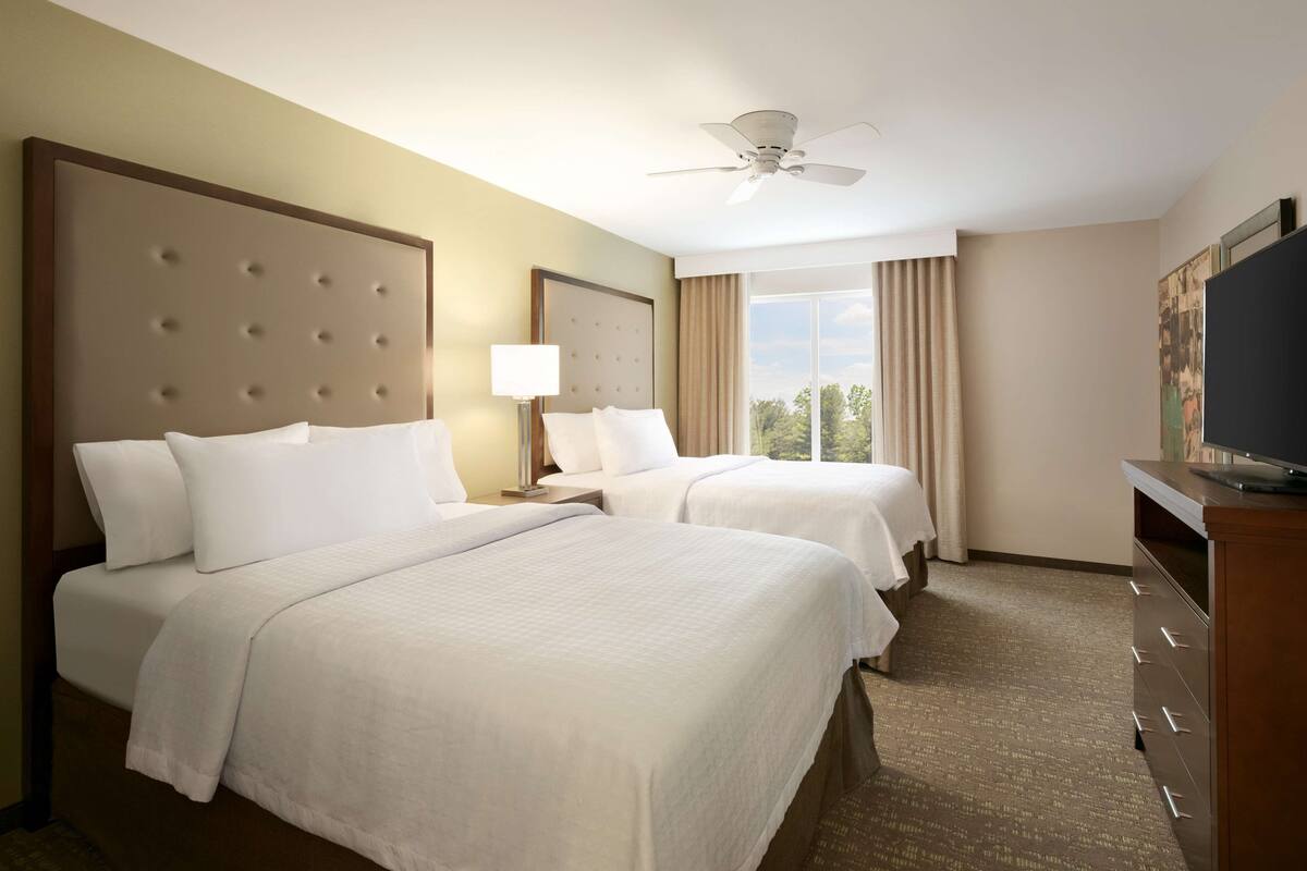 Een hotelkamer met twee bedden, een plafondventilator, een televisie en een groot raam met gordijnen.