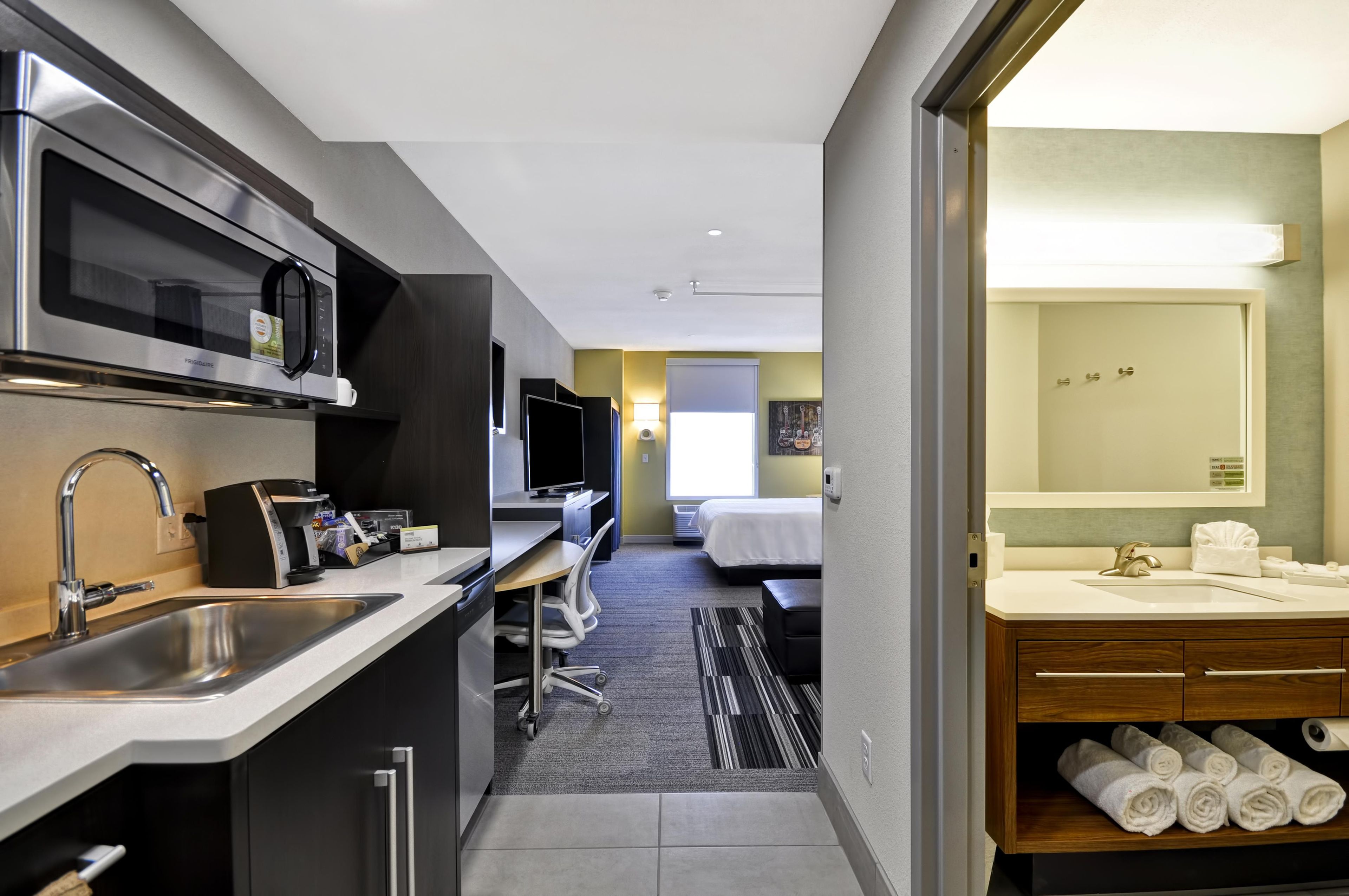A compact hotel room with a kitchenette, a bed, a bathroom, and a view of the city.