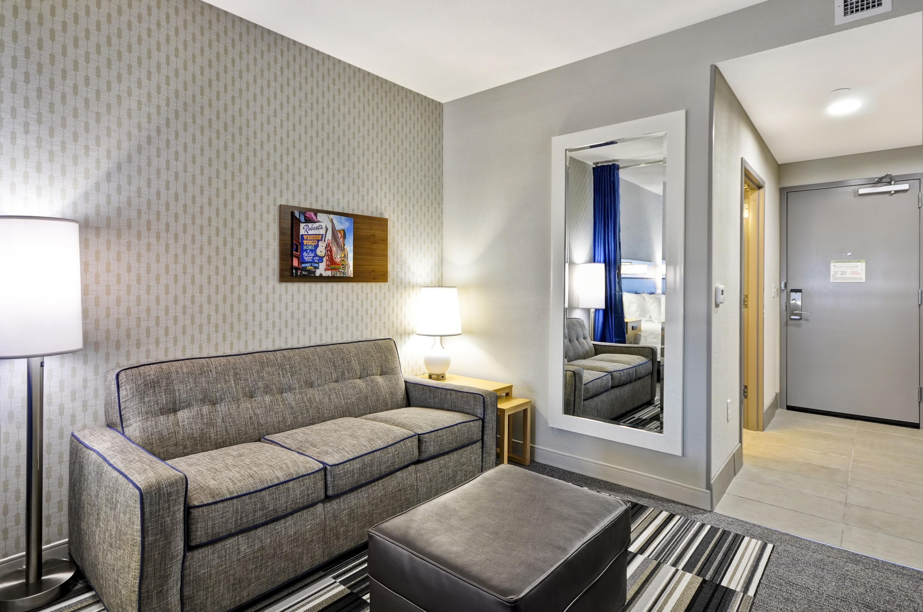 A hotel room with a sofa, a small table with a lamp, a mirror, and a door leading to another room.