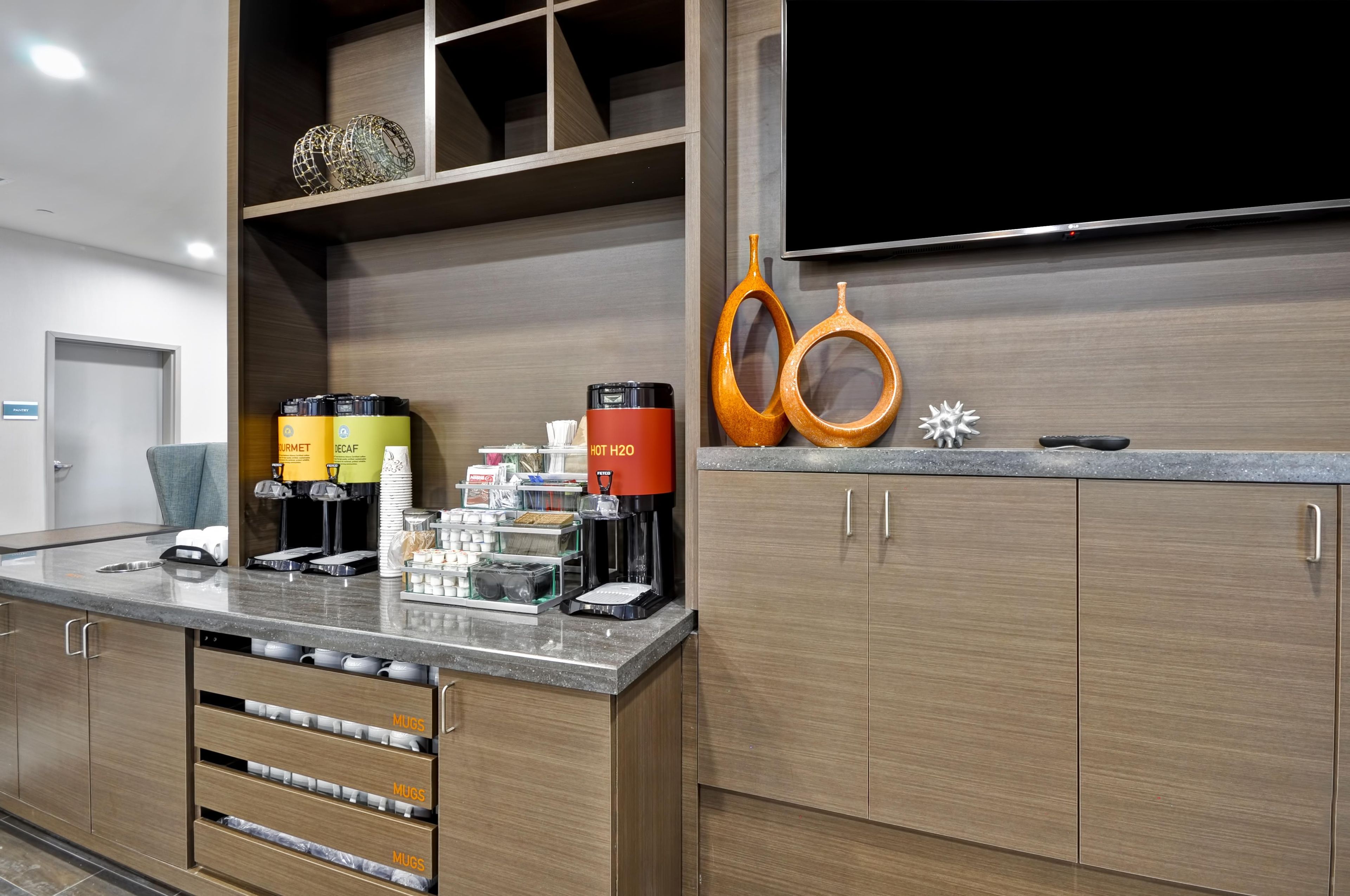 Una camera d'albergo con angolo cottura, dotata di macchina del caffè, dispenser per tè e dispenser di acqua calda. Sotto il bancone si trovano pensili e un televisore montato a parete.