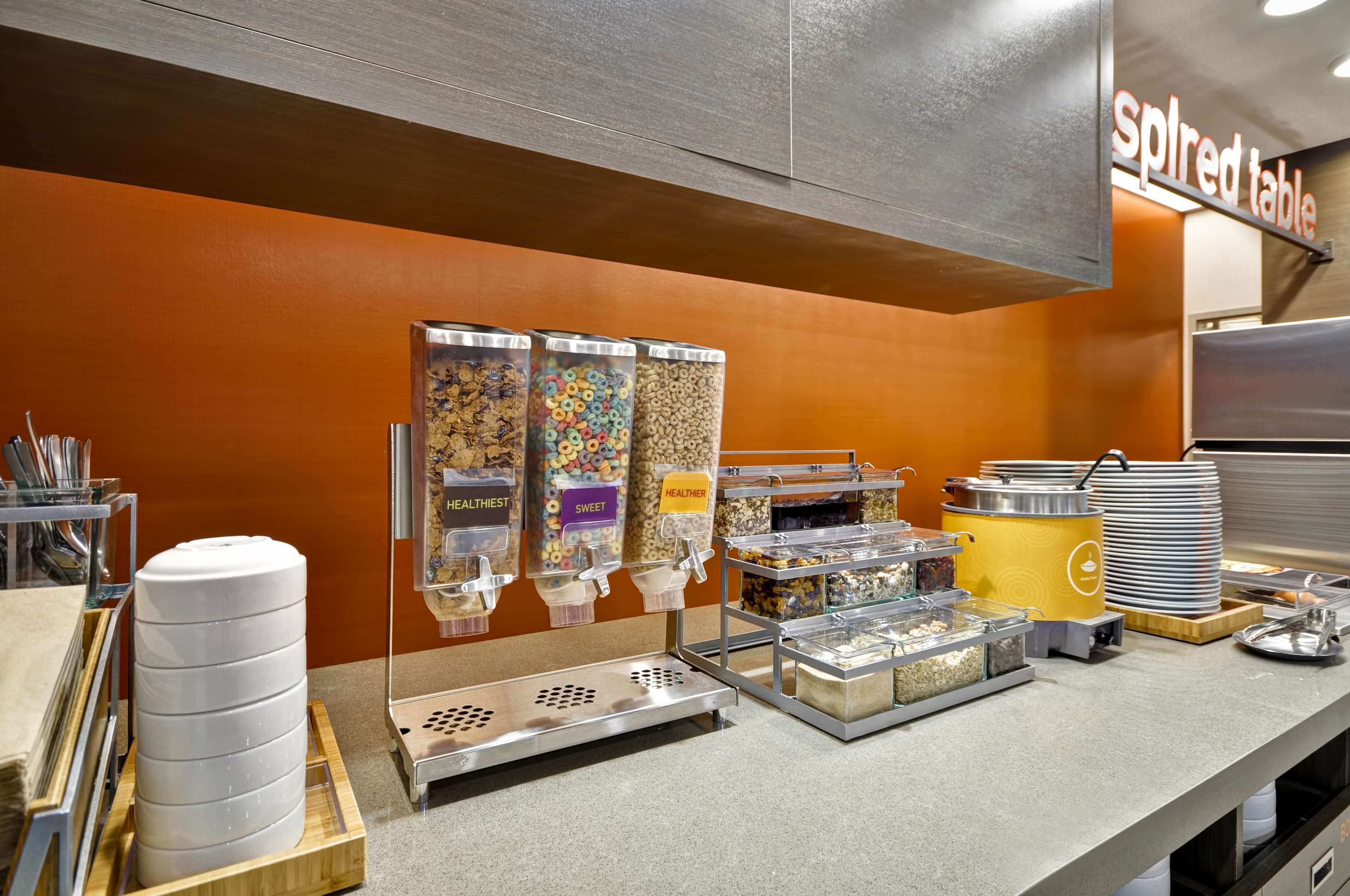 Un área de buffet con dispensadores etiquetados para cereales, una pila de platos y un recipiente amarillo.