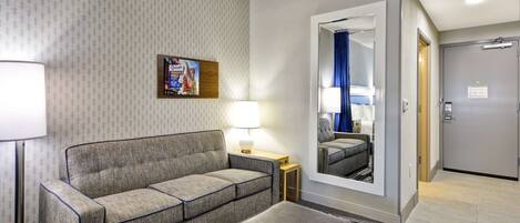Ein Hotelzimmer mit grauem Sofa, einem kleinen Tisch mit Lampe, einem Spiegel und einer Tür rechts.