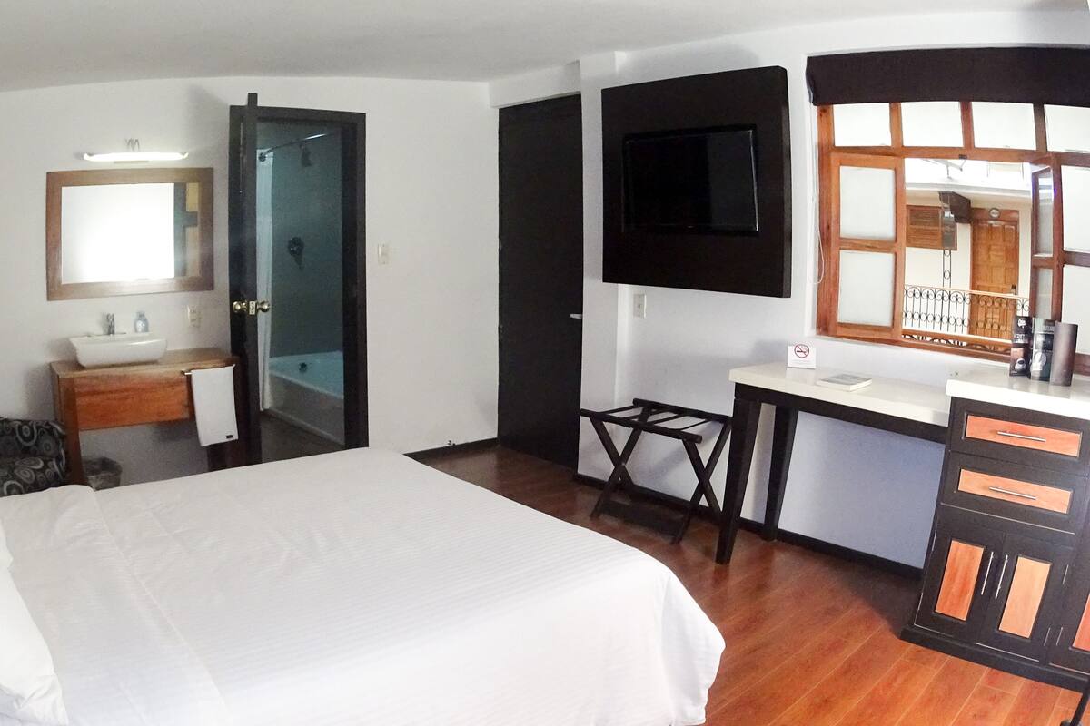 Habitación de hotel con cama, escritorio, silla, baño con lavamanos y espejo, y televisor montado en la pared.