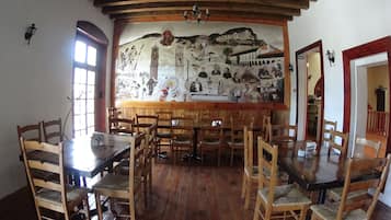 Un comedor con mesas y sillas de madera, un mural en la pared que representa escenas históricas y una araña.