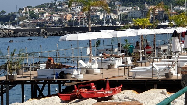 A waterfront restaurant with outdoor seating, a red boat, and a cityscape in the background. - Light vacation apartment in Forio (Isola d 'Ischia - NA) (Casamicciola Terme)