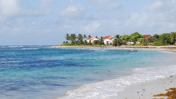 Una playa con agua cristalina azul, arena blanca y casas con tejados rojos.