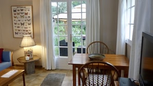 Een woonkamer met een bank, een tafel met stoel, een lamp, een televisie en een raam met gordijnen.