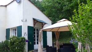 Een overdekt zitgedeelte met zwarte rieten meubels, een wit gebouw met groene luiken en een tuin met diverse planten.