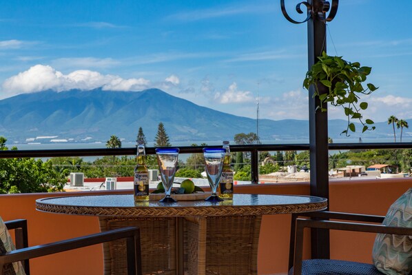 Una terraza con vista a la montaña, una mesa con dos vasos y una botella, y mobiliario de mimbre.