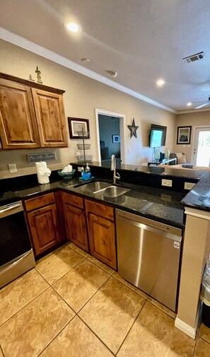 A kitchen with wooden cabinets, a stainless steel dishwasher, a black countertop, and a tiled floor.
