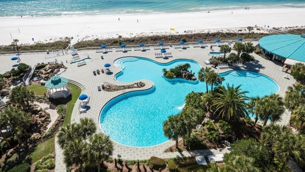 A resort with a large, winding pool surrounded by palm trees and lounge chairs, adjacent to a beach.