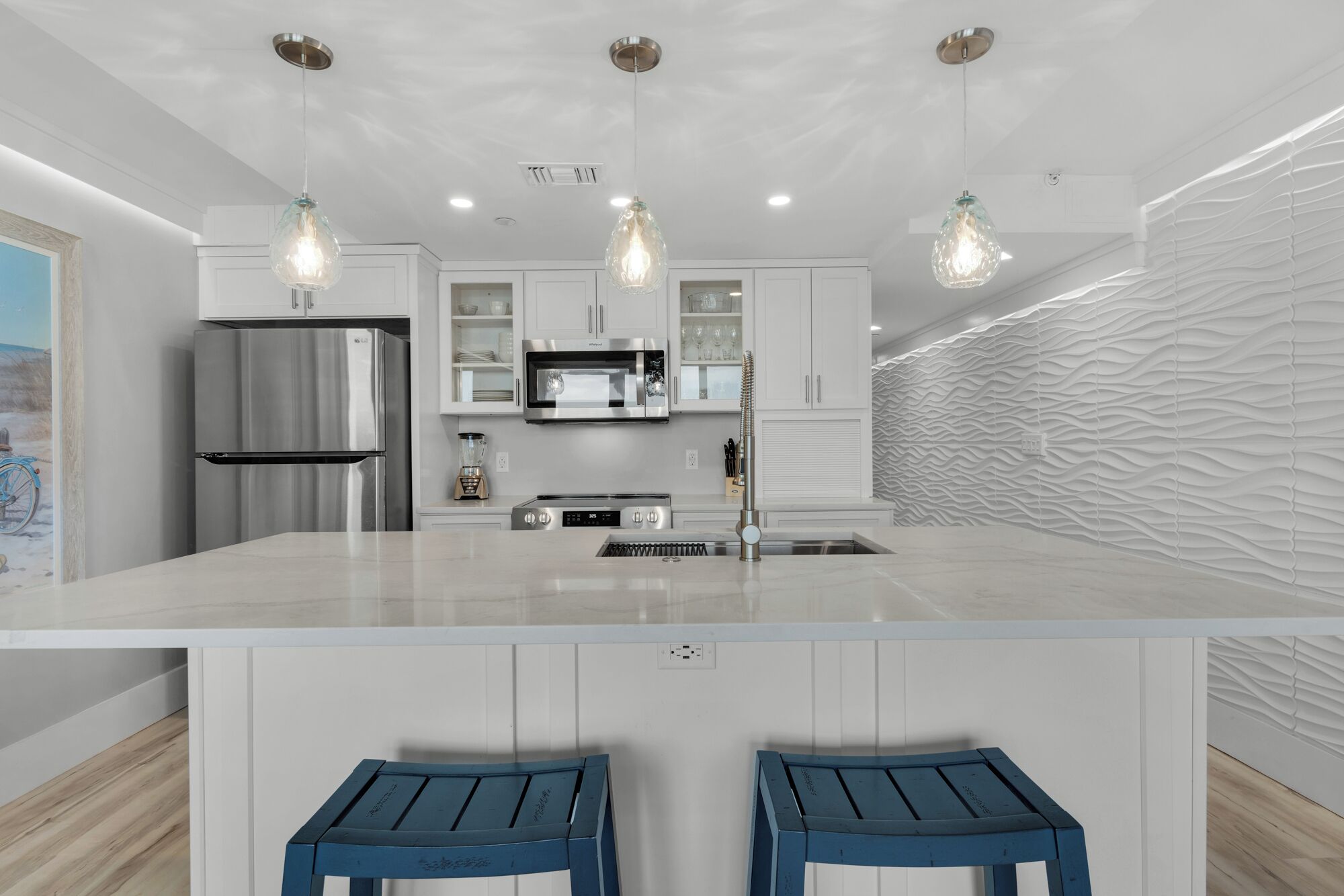 Een moderne keuken met een centraal eiland, roestvrijstalen apparatuur, hanglampen en een gebobbelde achterwand.