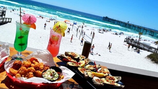 Blick auf einen Strand mit einem Tisch voller Speisen und Getränke, darunter frittierte Gerichte und Meeresfrüchte, mit Blick auf das Meer und einen Steg.