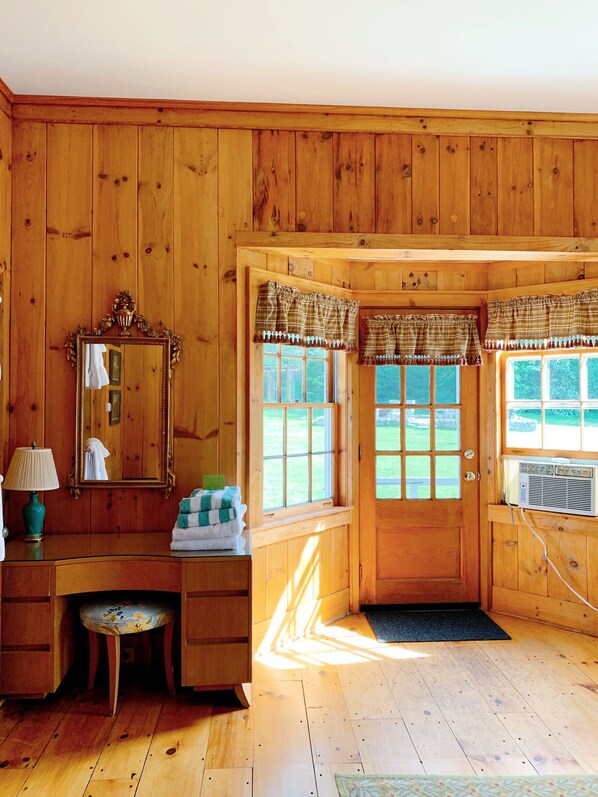 L’intérieur d’un chalet en bois, avec une coiffeuse, un miroir et deux fenêtres ornées de rideaux à carreaux.