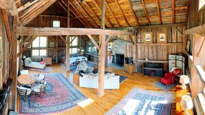 A rustic cabin interior with wooden beams, a fireplace, a dining area, and a living room with a sofa and chairs.