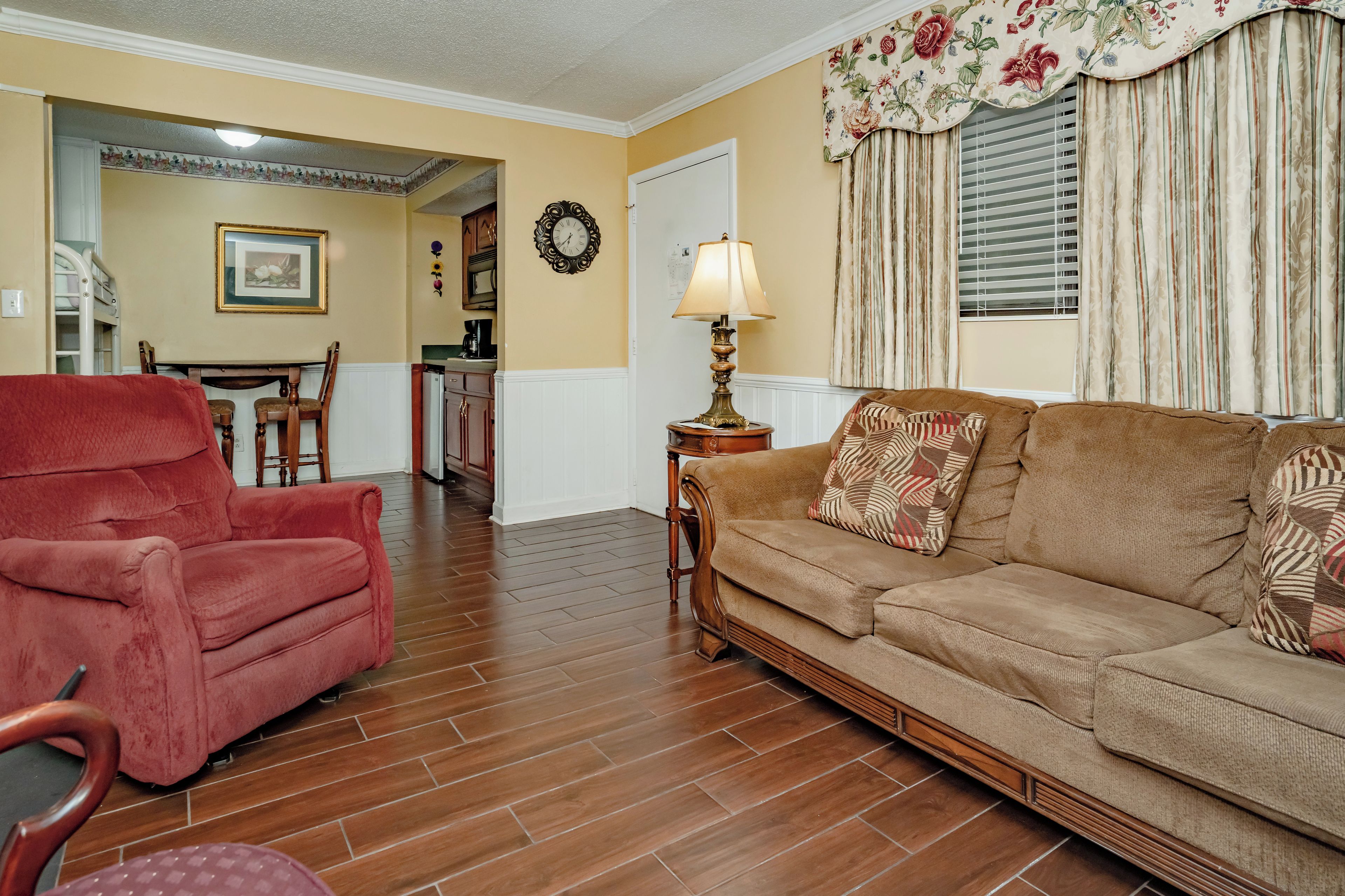 A living room with a red armchair, a beige sofa with patterned cushions, wooden flooring, a dining area with a table and chairs, and a kitchen in the background.