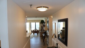 A hallway with a dining area in the background, a mirror on the wall, and a lamp on a console table. - Surf Dweller 611 by Alicia Hollis Rentals FREE TICKETS & BEACH SERVICE! (Fort Walton Beach)