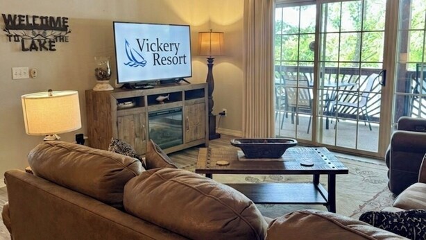 A living room with a ceiling fan, a TV displaying 'Vickery Resort,' a sofa, a coffee table, and a view of the outdoors through large windows. - Enjoy sunsets, lake activies and Sports TV pkg at this Vickery Resort Condo (Hollister)