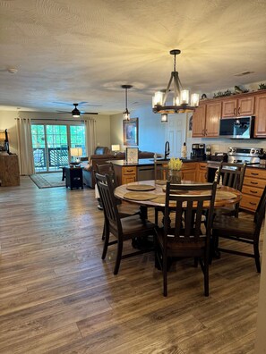 A spacious living area with a dining table, wooden chairs, a kitchen with modern appliances, and a ceiling fan.