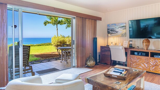 A modern living room with a glass door leading to a balcony with a view of the ocean. - Ground Floor Condo with Ocean and Mount Makana Views with A/C (Princeville)