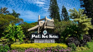 A sign for 'Pali Ke Kua Hideaways Pizza Pub' with a wooden structure and lush greenery around. - Ground Floor Condo with Ocean and Mount Makana Views with A/C (Princeville)