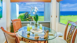 A glass-top table with chairs, a vase with flowers, and a view of the ocean through large windows with blinds. - Ground Floor Condo with Ocean and Mount Makana Views with A/C (Princeville)