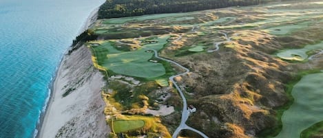 Vue aérienne d’un parcours de golf situé sur la côte, avec une plage, des fairways verdoyants et des bunkers de sable.