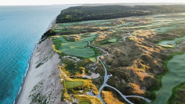 An aerial view of a coastal golf course with a beach, green fairways, and sand bunkers.
