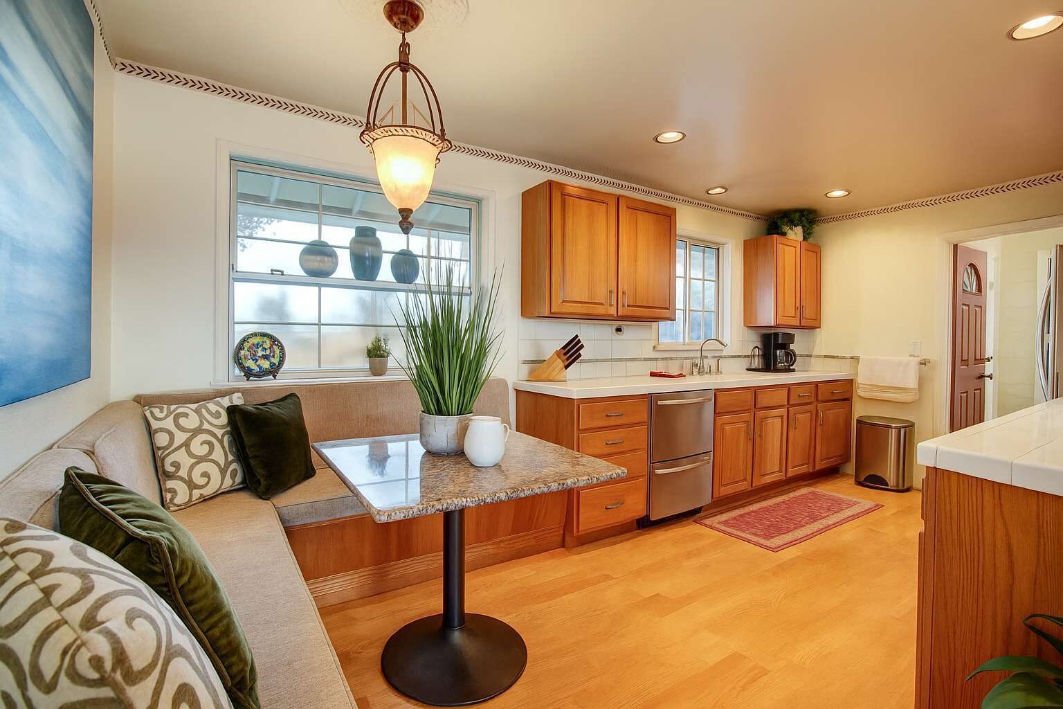 A modern kitchen with wooden cabinets, a stainless steel refrigerator, and a built-in oven. There is a dining area with a table and chairs, and a large window providing natural light.