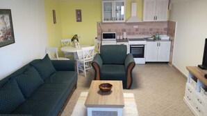 Una sala de estar con un sofá verde, un sillón estampado y una mesa de centro de madera. Hay una pequeña mesa con sillas y un jarrón de flores. En el fondo, la cocina tiene armarios blancos, un microondas y una estufa.