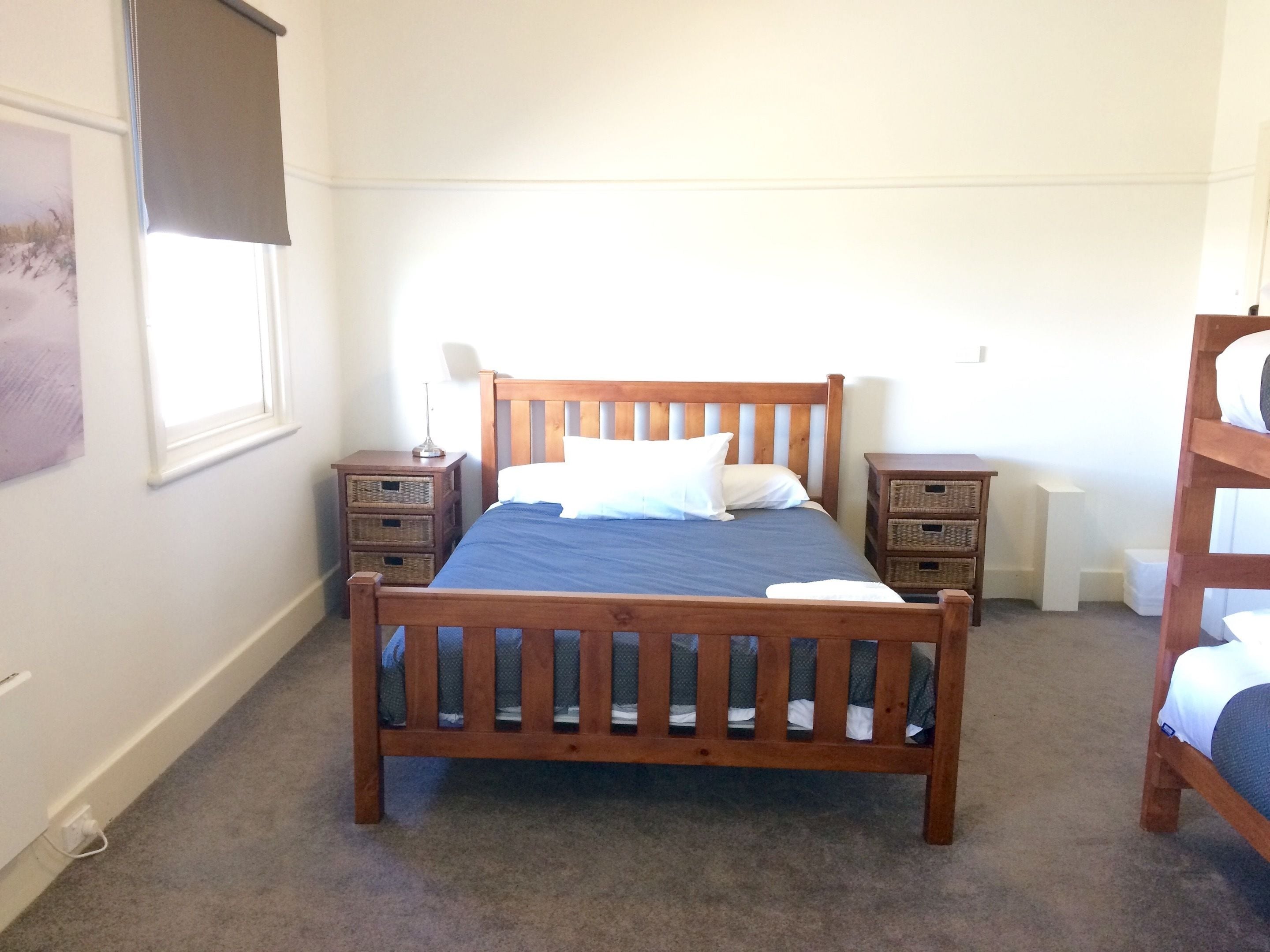 A bedroom with a wooden bed, two bedside tables, a window with a blind, and a bunk bed in the corner.