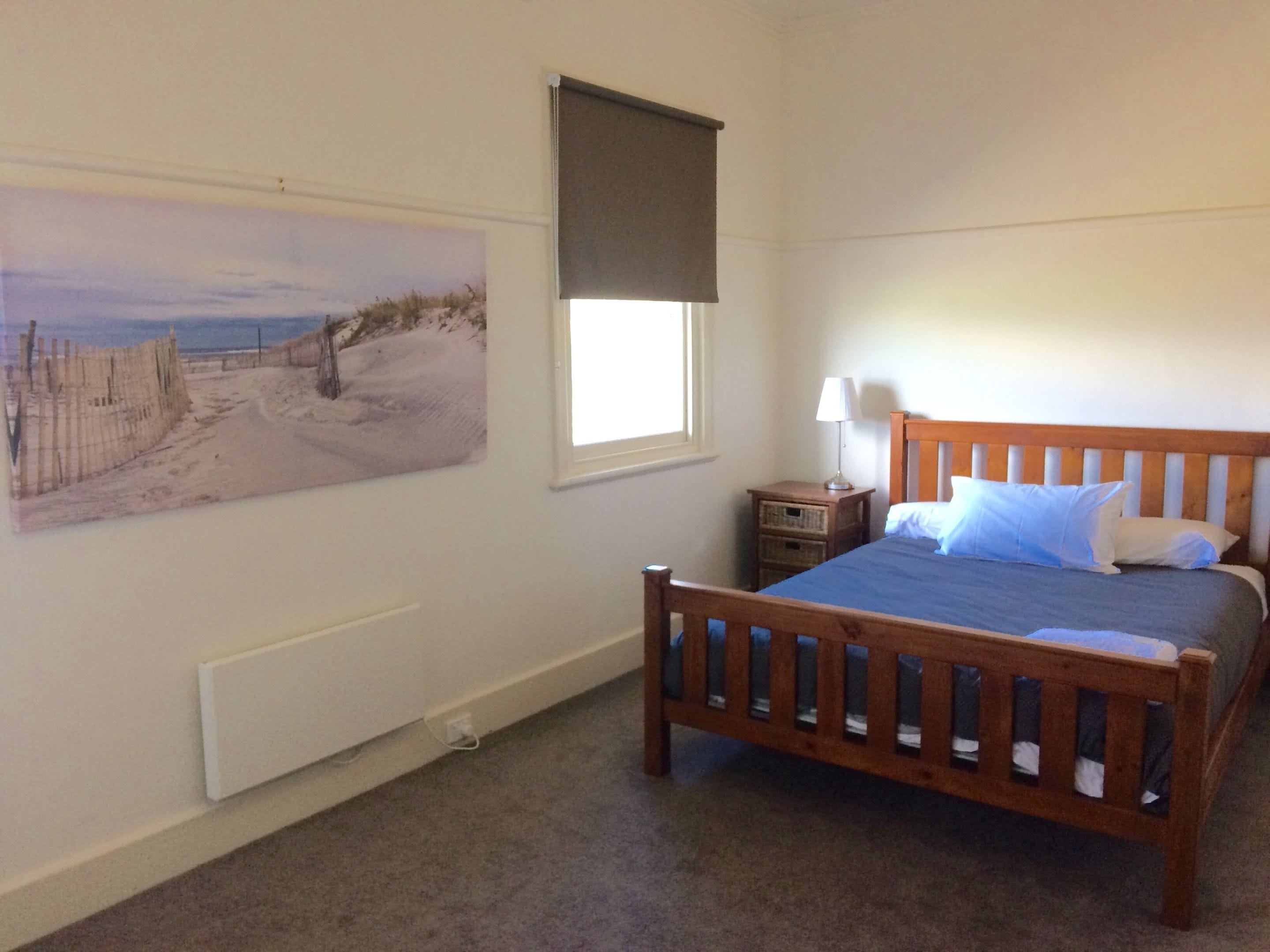 A bedroom with a wooden bed, a nightstand, a lamp, a window with a blind, and a beach-themed painting on the wall.