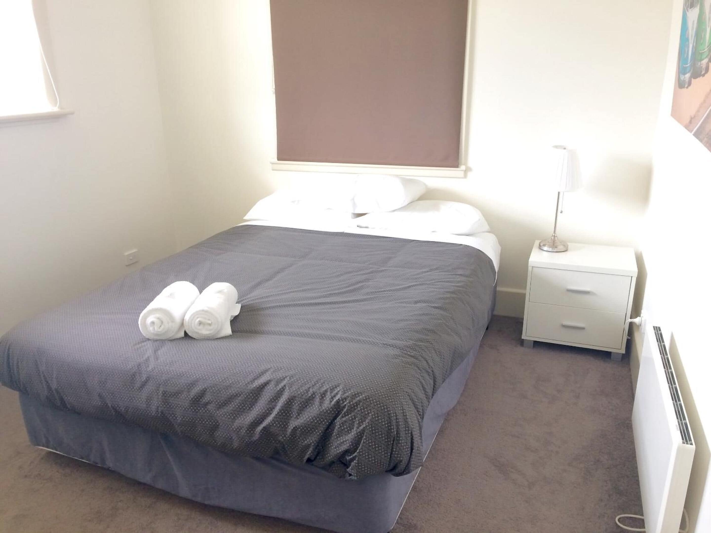 Un lit bien fait, recouvert d’une couverture grise, avec deux serviettes enroulées, une table de chevet avec une lampe et un radiateur fixé au mur.
