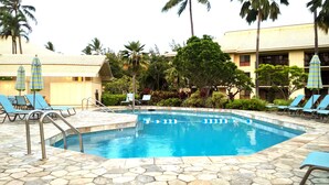 A swimming pool with lounge chairs and umbrellas, surrounded by palm trees and a building with a tiled roof.