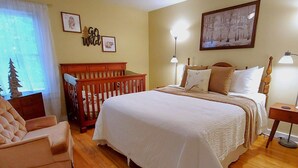 A bedroom with a large bed, a wooden crib, a chair, and a nightstand with a lamp.