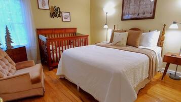 Un dormitorio con una cama grande, una cuna de madera, una silla y una mesita de noche con una lámpara.