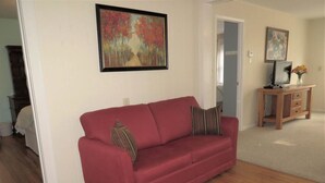 A red sofa with two pillows, a wooden cabinet with a laptop, and a painting on the wall.