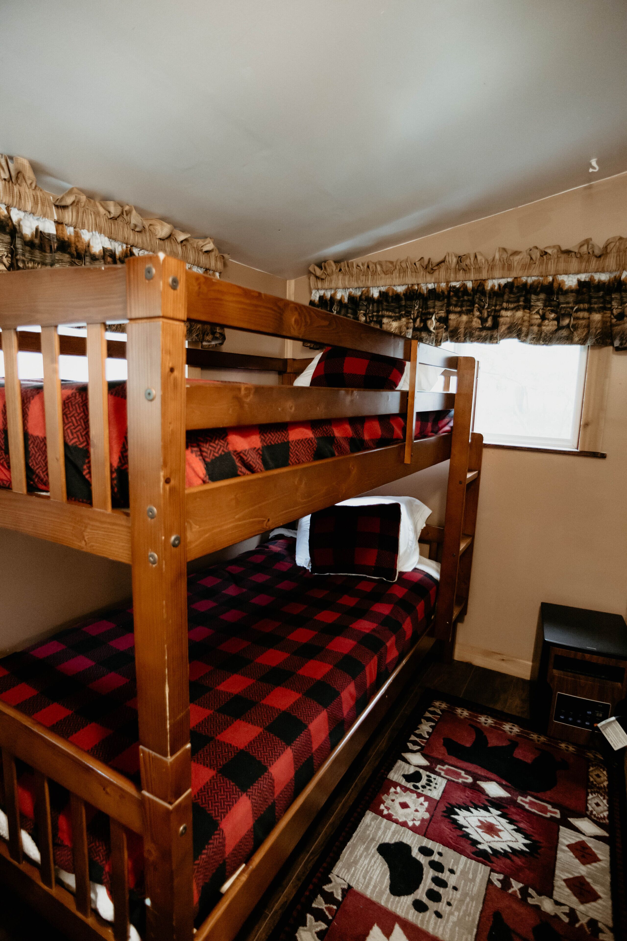 A bunk bed with a checkered bedspread, a window with curtains, and a patterned rug.