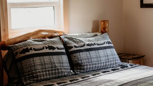 A bedroom with a wooden headboard, a window with a curtain, a bedside table with a lamp, and a bed with patterned bedding. - Cozy Creekside Cottage  (Custer)
